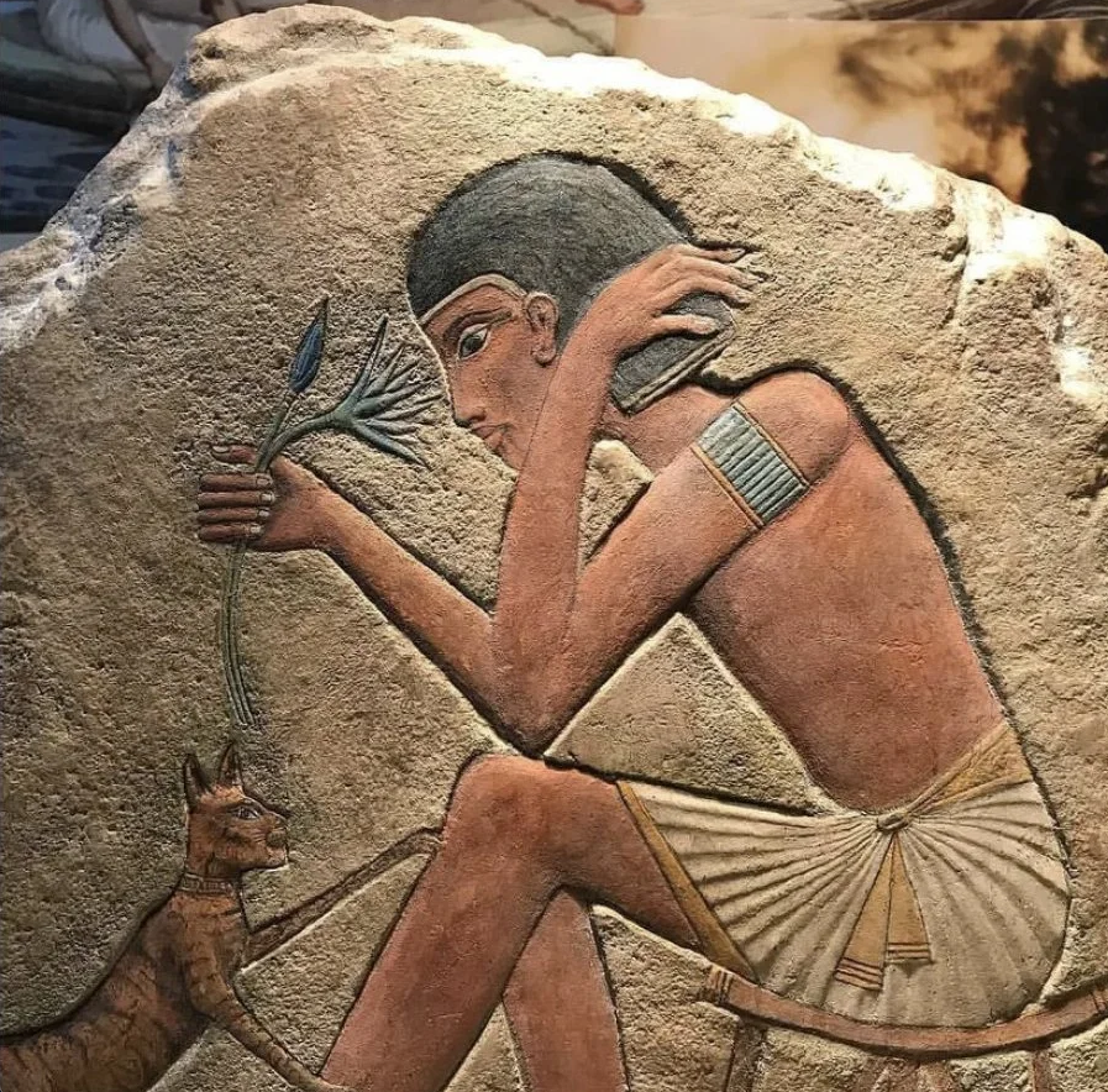 Ancient Egyptian stone relief depicting a figure holding a blue lotus flower with a cat at their feet